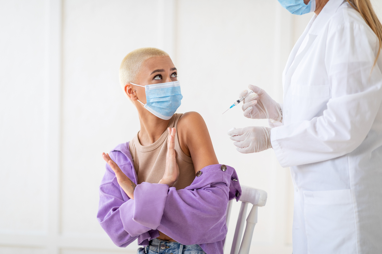 A woman with short blonde hair in a pink shirt wearing a face mask refuses a shot from a woman in a lab coat and face mask.